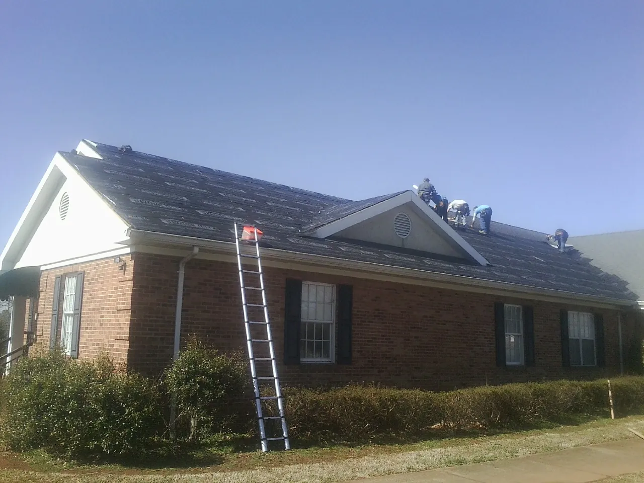 Crew tearing shingles off a brick ranch roof with a ladder set — Emergency Roof Repair work by our crew in Bancroft