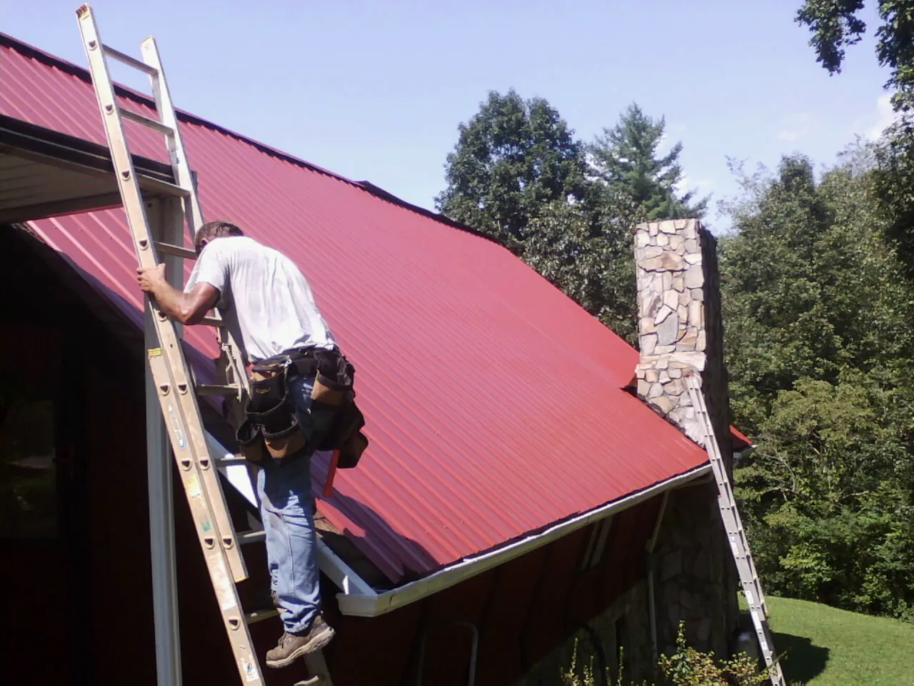 Roofer on a ladder beside a red metal roof and stone chimney — Soffit & Fascia Repair by Roofing Brothers in Bancroft